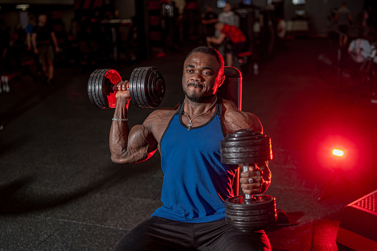 male afroamerican athlete is doing biceps dumbbells curls while sittiing gym sport chair